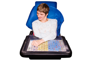 A person seated in a blue chair, using a communication device with symbols on a touchscreen.